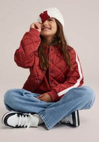 Raudona siuvinėta striukė su baltais dryžiais, raudonas džemperis, šviesiai mėlynos spalvos džinsai ir juodi balti sportbačiai. Balta kepurė su raudonu „M“ akcentu.