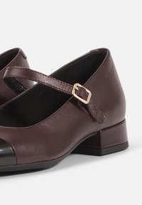 Sapatos Mary Jane de couro borgonha, com biqueira preta, tira ajustável e salto baixo em bloco. Textura lisa e design minimalista.
