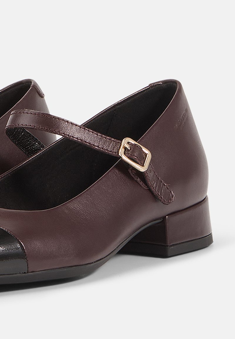 Burgunderfärgade läder Mary Jane-skor med svart tådyra, justerbar rem och låg blockklack. Slät yta och minimalistisk design.