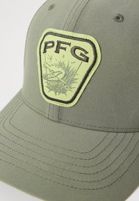 Cască verde din material textil, cu un design brodat. Previne o etichetă galbenă și neagră, cu un grafic al unui aligator și motive vegetale.