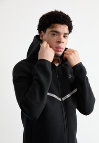 Sweatshirt com fecho éclair preta feita de tecido suave, com um design de risca contrastante branca ao longo do peito e capuz com cordão.