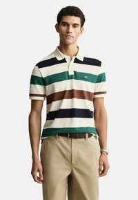 Gestreepte polo in crème, met groene, marineblauwe, bruine en witte horizontale strepen. Korte mouwen en een klassieke kraag. De broek is kaki.