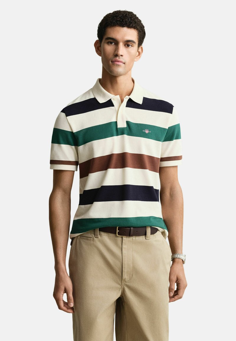 Gestreepte polo in crème, met groene, marineblauwe, bruine en witte horizontale strepen. Korte mouwen en een klassieke kraag. De broek is kaki.