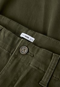 Olivengrønne bukser med en glatt tekstur i stoffet, med knappelukking og en merkelapp som viser "TWO SOON" med størrelse 32/32.