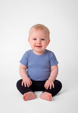 Bambino sorridente con capelli chiari seduto a gambe incrociate su una superficie bianca, indossa una maglietta blu a maniche corte e pantaloni neri.
