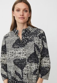 Camicia a maniche lunghe color crema chiaro con un motivo astratto nero. Caratterizzata da un colletto e maniche a tre quarti, realizzata in un tessuto testurizzato.