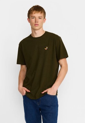 Jonge man draagt een donkergroen T-shirt met een klein kleurrijk geborduurd logo, blauwe spijkerbroek, staat met handen in de zakken tegen een witte achtergrond.