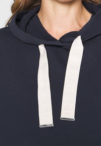 Mörkblå huvtröja med justerbar dragsko som har krämfärgade, logotryckta snören och silverfärgade metalldelar. Slät tygkvalitet.