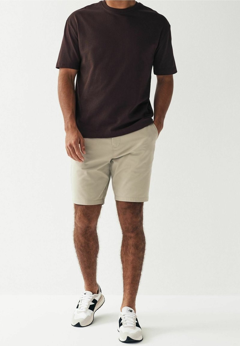 Homme portant une chemise à manches courtes marron foncé, un short beige et des baskets blanches avec des accents noirs, debout devant un fond clair uni.