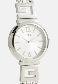 Ρολόι ασημί απόχρωσης με στρογγυλή κάσα, λευκό καντράν, κρυστάλλινους δείκτες, υφή στα σημεία, και βραχιόλι από αλυσίδα με λεπτομέρειες λογότυπου.