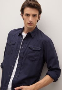 Camisa azul marino con botones, dos bolsillos delanteros, cuello de solapa y mangas remangadas. Hecha de tela suave con una textura lisa.