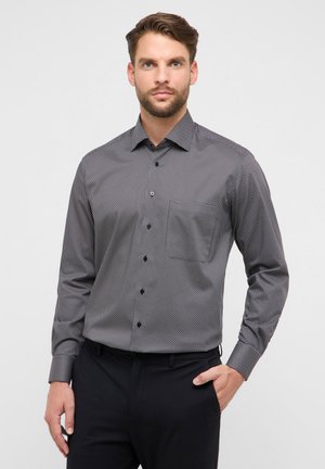 Mann trägt ein eng anliegendes, langärmeliges Hemd mit kleinem weißen Punktmuster und schwarze Hose, steht vor hellem, einfarbigem Hintergrund.