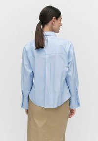 Jasnoniebieska koszula z długimi rękawami, z plisą z tyłu, zaokrąglonym brzegiem i mankietami z guzikami. Nosi na sobie beżową spódnicę.