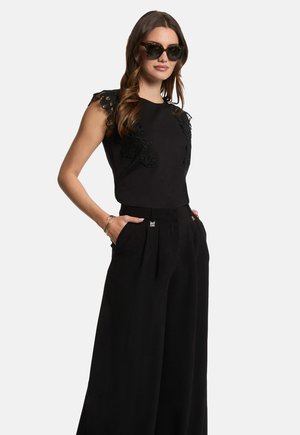 Black sleeveless top with lace detail at shoulders, paired with high-waisted black wide-leg trousers. Model wears patterned sunglasses.