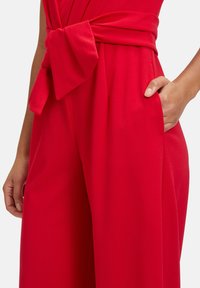 Mono rojo con una cinta atada en la cintura, bolsillos y una textura de tela suave y caída. El diseño destaca un ajuste holgado y una silueta elegante.