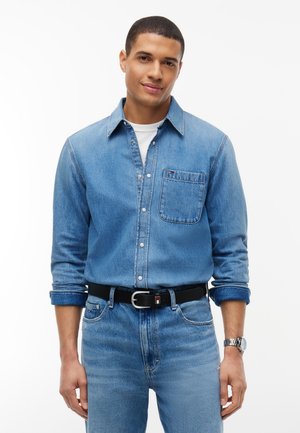 Mand iført en lyseblå denimskjorte og jeans med opkrogede ærmer, et sort bælte og et sølv ur, står foran en ensfarvet baggrund.
