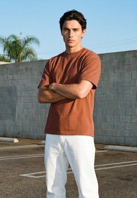 Młody mężczyzna o ciemnych włosach stoi na parkingu z założonymi skrzyżowanymi ramionami, ubrany w brązowy t-shirt i białe spodnie, na tle szarej ściany z bloków.