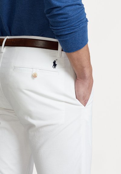 Valkoiset puuvillapantsit, joissa on takatasku ja pieni laivaston sininen logo. Ruskea nahkavyö kontrastoi kangasta. Sininen neule yllä.