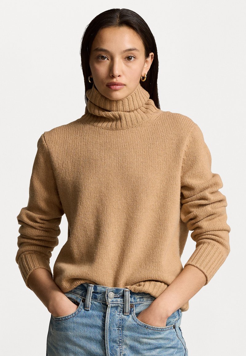 Beige gebreide trui met een hoge ribgebreide col, met lange mouwen en een relaxte pasvorm. Gekocht met lichtblauwe denim, met handen in de zakken.