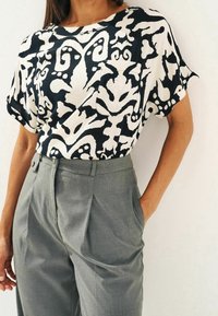 Schwarze und weiße gemusterte Kurzarmbluse mit einer Tasche, kombiniert mit hochgeschnittenen grauen Hosen mit Falten und seitlichen Taschen.