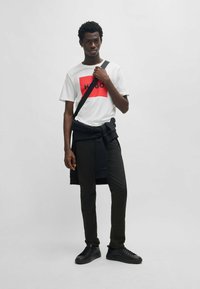 T-shirt blanche avec un graphique rectangulaire rouge et des lettres noires, associée à un pantalon noir et des baskets noires. Comprend un sac bandoulière noir.