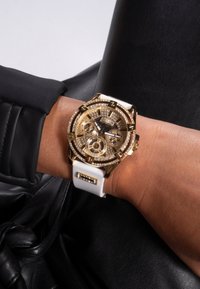 Orologio color oro con cinturino in gomma bianca, accenti di cristallo e quadrante testurizzato con tre sotto-quadranti e marcatori delle ore audaci.