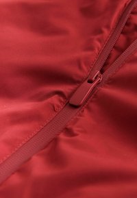 Detail zipsovaného červeného textilu, s hladkou textúrou a leskom. Zipper je v rovnakej červenej farbe, integrovaný do švu.