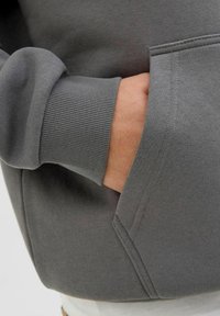 Szary sweatshirt z prążkowanymi mankietami i kieszenią kangurką; gładka tekstura materiału; zbliżenie pokazujące detal kieszeni i fason mankietu.