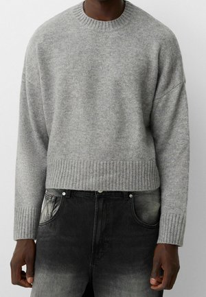 Homme portant un pull gris clair en maille côtelée à col rond et un jean noir délavé, visible du cou aux hanches sur un fond uni.