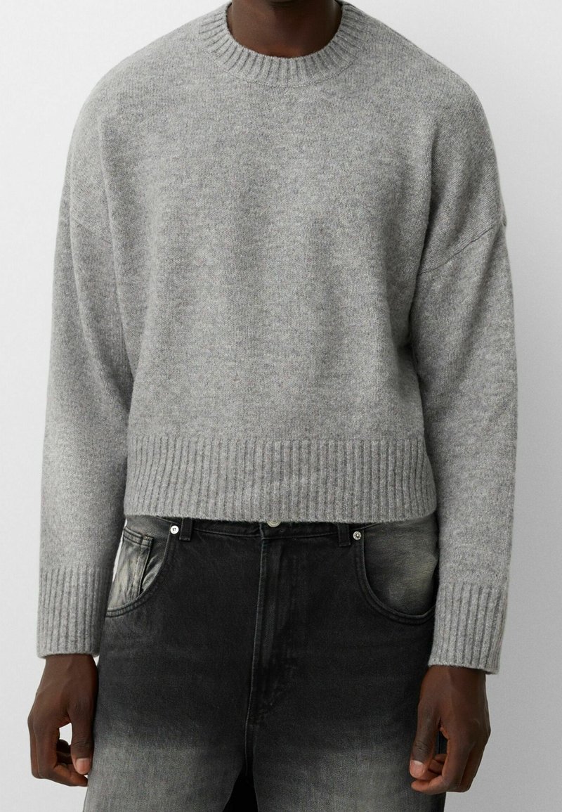 Homme portant un pull gris clair en maille côtelée à col rond et un jean noir délavé, visible du cou aux hanches sur un fond uni.