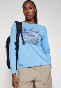 Long-sleeve, light blue shirt with a graphic print in purple tones. Paired with light tan pants and a black backpack.