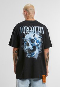 Fekete, bő szabású póló, amelyen egy kék koponya grafika látható tüskékkel, felette pedig kiemelt betűkkel a "ELFELEJTETT" szó olvasható. Narancssárga címke az alján.
