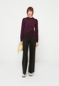 Lila Pullover und schwarze weit geschnittene Hosen, dazu eine leichte goldene Tasche. Das Schuhwerk ist hellgrau mit schwarzen Sohlen. Glatte Textur und figurbetontes Design.