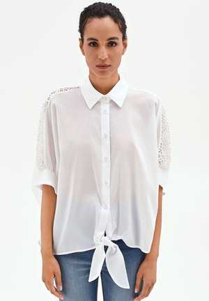 Femme portant une chemise blanche boutonnée avec des détails en dentelle sur les manches, nouée à la taille, associée à un jean bleu sur fond uni.