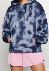 Felpa tie-dye in tonalità di navy e grigio con tasca frontale e cordini. Indossata sopra un paio di pantaloni corti sportivi rosa, con un patch del logo.