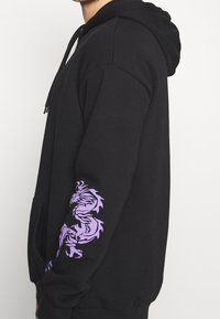 Persona che indossa un hoodie nero con una grafica di un drago viola sulla manica destra, in piedi di lato su uno sfondo bianco semplice.
