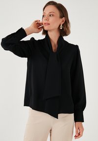 Black blouse with a neck tie, puffed sleeves, buttoned cuffs, and smooth texture. Worn with beige pants against a neutral background.