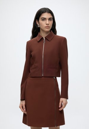 Femme aux longs cheveux foncés porte une veste marron zippée et une jupe assortie avec une fermeture éclair sur le côté, debout devant un fond clair uni.