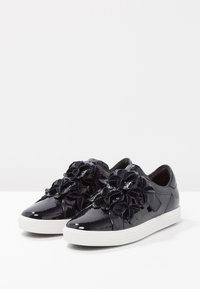 Baskets en cuir verni marine présentant un ornement floral texturé à l'avant, avec une semelle en caoutchouc blanche et un design épuré.