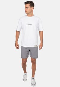 T-shirt blanc à manches courtes avec un logo script noir, associé à un short gris avec broderie, et des baskets blanches. Design simple.