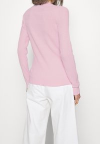 Kvinna klädd i en ljusrosa texturerad stickad tröja och lösa vita byxor, fotograferad bakifrån mot en vit bakgrund.