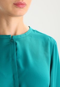 Blusa de color aguamarina, fabricada en tejido suave, con escote redondeado, que presenta un pequeño cierre de botón en el cuello y un diseño sencillo.