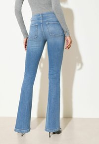 Frau trägt hoch taillierte, hellblaue Schlagjeans und graues langärmeliges Oberteil, steht vor einer schlichten weißen Wand.