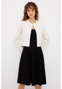Cardigan corto bianco con motivo testurizzato sul giromanica, abbinato a un vestito A-line senza maniche di colore nero. Design semplice ed elegante.