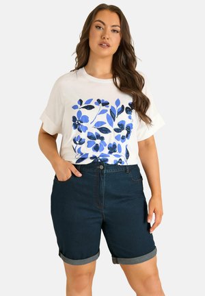 Frau trägt ein weißes T-Shirt mit blauem Blumenmuster und dunkelblaue Jeansshorts, hochgekrempelt, steht mit einer Hand in der Tasche.