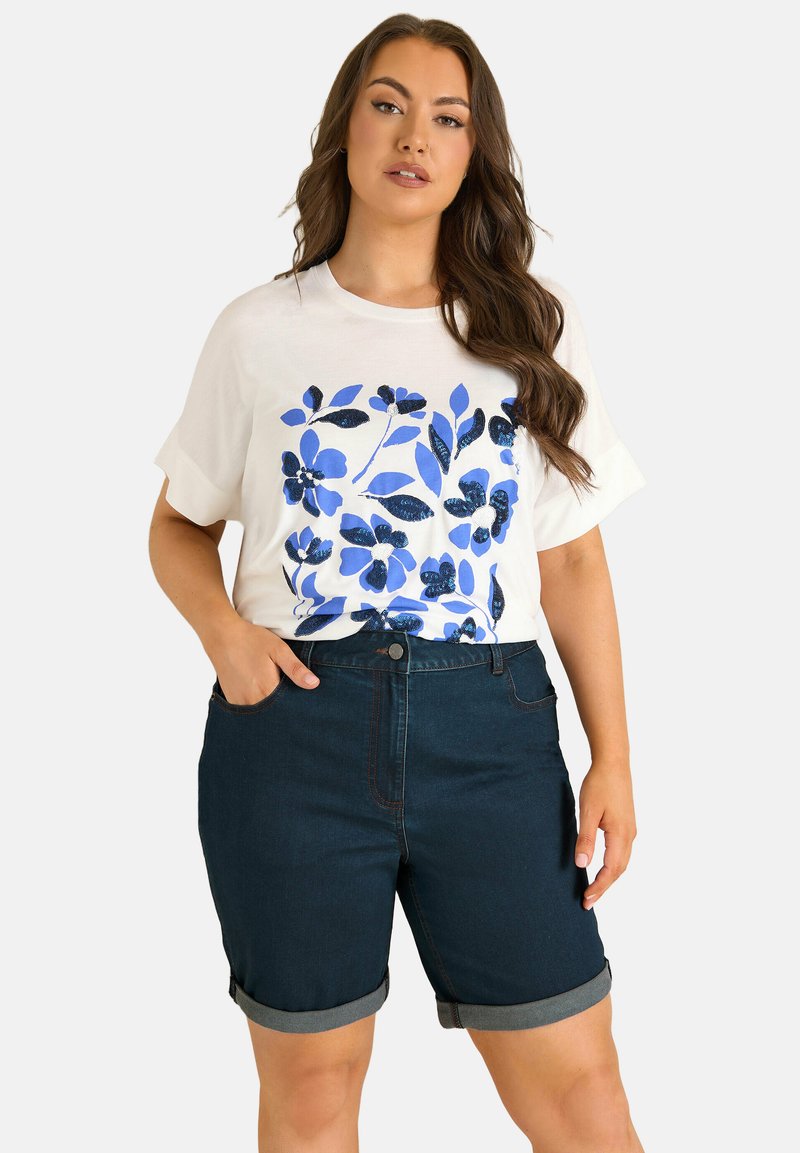 Woman wearing a white t-shirt with blue floral print and dark denim rolled-up shorts, standing with one hand in pocket.