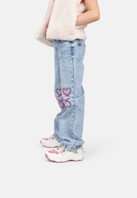 Jeans in denim blu chiaro con ricami a forma di cuore viola sui polpacci, abbinati a sneakers bianche e rosa. Il modello indossa un gilet morbido.