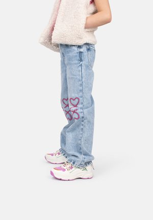 Un jean en denim bleu clair avec des broderies de cœurs violets sur les jambes, associé à des baskets blanches et roses. Le modèle porte un gilet pelucheux.