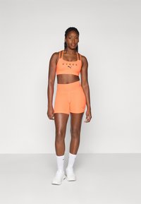 Oransje sports-BH med tynne stropper og svart logo, sammen med matchende oransje shortser. Modellen har på seg hvite joggesko og ankellange hvite sokker.