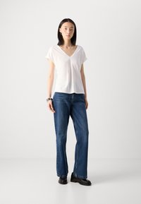 T-shirt blanc à col en V avec des manches courtes, associé à un jean bleu droit et des chaussures noires à plateforme, se tenant sur un fond neutre.